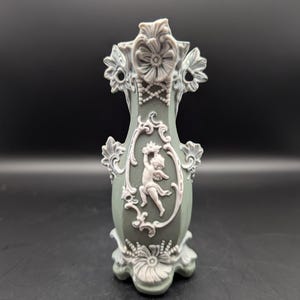 May include: A sage green decorative vase with white floral and cherub accents. The vase features a detailed relief design with a cherub in an oval frame. The top and base have ornate floral patterns, and the vase is approximately 18 cm tall.
