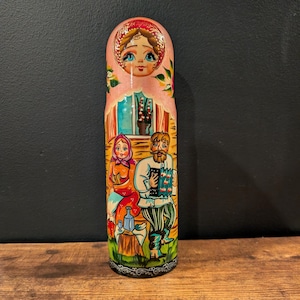 May include: A hand-painted Russian nesting doll, featuring a pink exterior with a painted scene of a man and woman. The doll has a detailed floral design and a black base. The figures are dressed in traditional clothing.