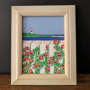 May include: A framed print featuring a coastal scene with a white picket fence adorned with red roses. A lighthouse stands on a green island under a blue sky. The frame is made of light-colored wood.