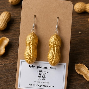 Könnte beinhalten: Ein Paar goldfarbene, erdnussförmige Ohrringe, die an einer braunen Pappkarte hängen. Die Karte zeigt den Text lyly_piezas_arte, eine Strichzeichnung von zwei Figuren, den Text Hecho a mano und den Instagram-Namen @lyly_piezas_arte.