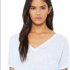 May include: A short-sleeved V-neck t-shirt in a light blue and white heathered fabric. The shirt features a relaxed fit with a simple neckline and short sleeves, paired with dark blue denim jeans. The material has a subtle speckled texture.