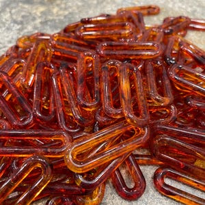 Op de afbeelding: Een verzameling doorschijnende, barnsteenkleurige plastic kettingschakels met een glanzende afwerking. Elke schakel is ovaal van vorm met afgeronde randen, wat een hoopje onderling verbonden onderdelen vormt in verschillende tinten oranje en bruin.