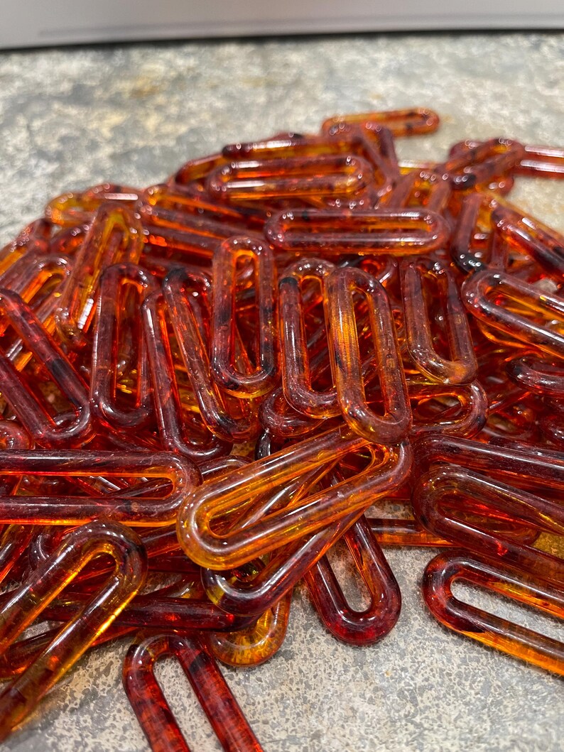 Op de afbeelding: Een verzameling doorschijnende, barnsteenkleurige plastic kettingschakels met een glanzende afwerking. Elke schakel is ovaal van vorm met afgeronde randen, wat een hoopje onderling verbonden onderdelen vormt in verschillende tinten oranje en bruin.