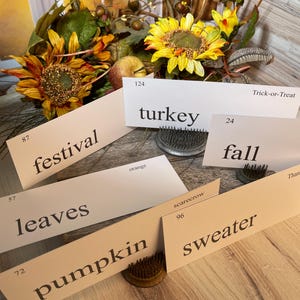 Op de afbeelding: Een verzameling rechthoekige witte kaarten met in het zwart gedrukte woorden zoals festival, turkey, trick-or-treat, fall, leaves, orange, scarecrow, pumpkin en sweater, elk met een klein nummer, gerangschikt bij zonnebloemen en herfstbladeren.
