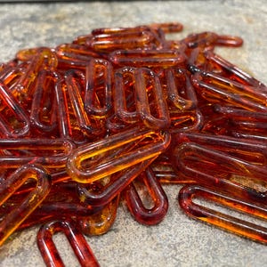 Op de afbeelding: Een verzameling doorschijnende, barnsteenkleurige plastic kettingschakels. Elke individuele schakel heeft een langwerpige ovale vorm met afgeronde randen en een gladde, glanzende afwerking. De schakels liggen op een hoop en vertonen verschillende tinten oranje en bruin.