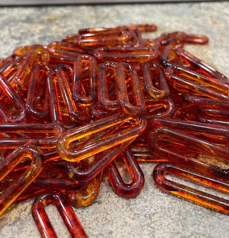 Op de afbeelding: Een verzameling doorschijnende, barnsteenkleurige plastic kettingschakels. Elke individuele schakel heeft een langwerpige ovale vorm met afgeronde randen en een gladde, glanzende afwerking. De schakels liggen op een hoop en vertonen verschillende tinten oranje en bruin.