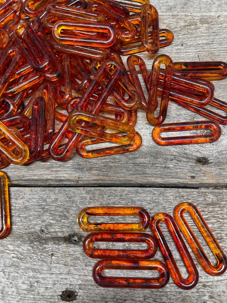 Op de afbeelding: Een verzameling doorschijnende amberkleurige rechthoekige plastic ringen met afgeronde hoeken. Deze langwerpige schakels hebben een gladde, glanzende afwerking en een hol midden, gerangschikt in een hoopje met enkele losse stukjes plat neergelegd.