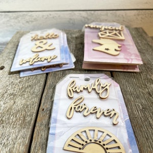 Op de afbeelding: Drie sets van lichtgekleurde, lasergesneden houten vormen en woorden in script, verpakt op rechthoekige kaarten. De kaarten hebben aquarelachtige achtergronden in blauw, roze en paars. De houten stukken bevatten teksten zoals hello & welcome, magical en family forever.