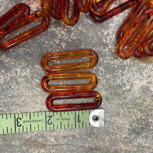 Op de afbeelding: Een verzameling van doorschijnende barnsteenkleurige ovale plastic kettingschakels. Een groen meetlint is langs de onderkant geplaatst, waaruit blijkt dat elke individuele schakel ongeveer 3,8 cm lang en 1,3 cm breed is.