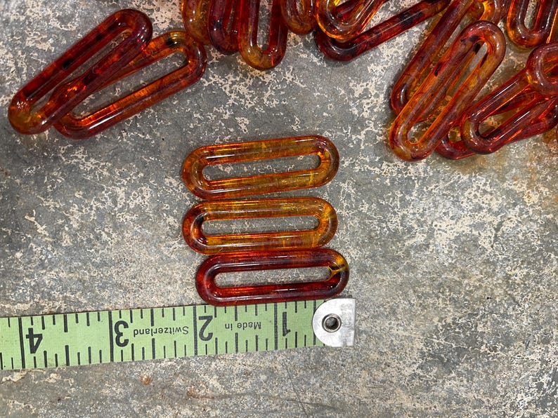 Op de afbeelding: Een verzameling van doorschijnende barnsteenkleurige ovale plastic kettingschakels. Een groen meetlint is langs de onderkant geplaatst, waaruit blijkt dat elke individuele schakel ongeveer 3,8 cm lang en 1,3 cm breed is.