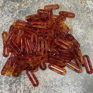 Op de afbeelding: Een grote verzameling doorschijnende, barnsteenkleurige plastic kettingschakels. Elke individuele schakel heeft een langwerpige ovale vorm met afgeronde randen en een gladde, glanzende afwerking. De schakels liggen verspreid op een hoop.