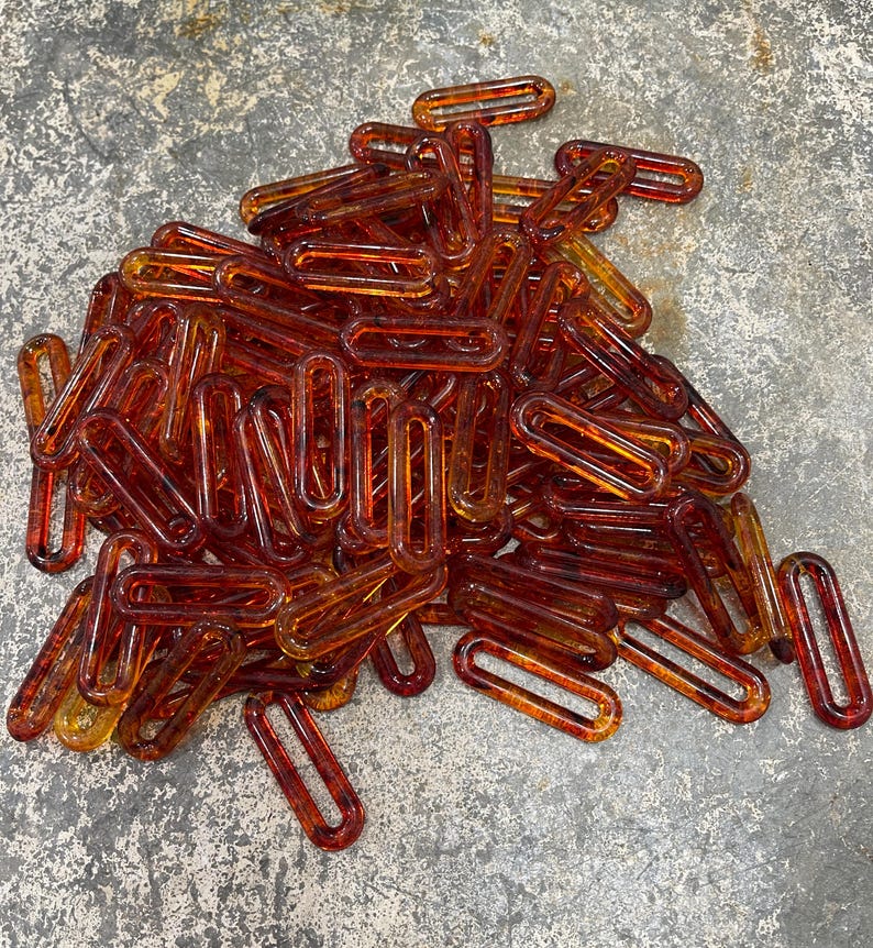 Op de afbeelding: Een grote verzameling doorschijnende, barnsteenkleurige plastic kettingschakels. Elke individuele schakel heeft een langwerpige ovale vorm met afgeronde randen en een gladde, glanzende afwerking. De schakels liggen verspreid op een hoop.