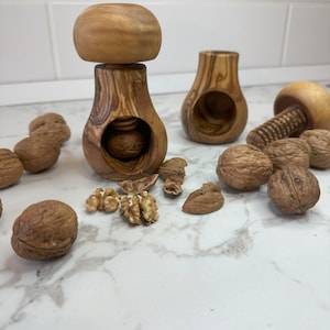 Könnte beinhalten: Holz-Nussknacker und Walnüsse liegen auf einer weißen Arbeitsplatte. Die Nussknacker haben einen pilzförmigen Kopf und einen zylindrischen Körper mit einem Loch zum Knacken von Nüssen. Walnüsse liegen verstreut um die Nussknacker, einige sind geöffnet.