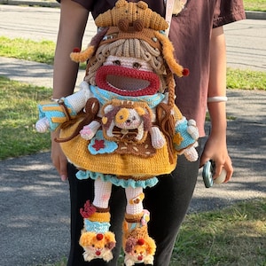May include: A handcrafted crochet backpack shaped like a doll with a croissant-style hat, brown hair, and a red mouth opening. The doll wears a yellow dress with blue accents, a small dog-shaped pocket, and matching crochet shoes with animal faces.