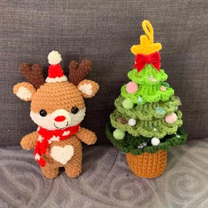 Könnte beinhalten: Gehäkelte Weihnachtsdekorationen mit einem Rentier und einem Weihnachtsbaum. Das Rentier hat einen rot-weißen Hut und Schal sowie ein weißes Herz auf der Brust. Der Baum ist grün mit bunten Ornamenten und einem gelben Stern.
