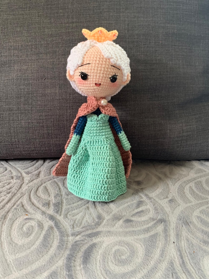 May include: A handmade crochet doll resembling a princess. The doll has a light peach skin tone, white hair, and a small gold crown. It wears a teal green dress, a pink cape, and a pearl necklace. The doll's face features black eyes and rosy cheeks.