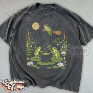 May include: A charcoal gray short-sleeve t-shirt featuring a graphic illustration of three green frogs in a pond setting. One frog flies on a broomstick above a cauldron, while two other frogs stir the pot amidst lily pads and cattails.