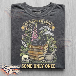 May include: A charcoal gray t-shirt featuring a graphic of a black cat sitting on a stack of books labeled Herbarium, Herbarium Maleficum, Alchemia Herbalis, De Venenis, and Liber Mortis, with a skull, mortar and pestle, and plants, plus the text All Plants Are Edible Some Only Once.