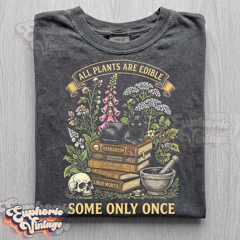May include: A charcoal gray t-shirt featuring a graphic of a black cat sitting on a stack of books labeled Herbarium, Herbarium Maleficum, Alchemia Herbalis, De Venenis, and Liber Mortis, with a skull, mortar and pestle, and plants, plus the text All Plants Are Edible Some Only Once.