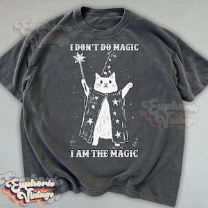 May include: A charcoal gray short-sleeve t-shirt featuring a white graphic of a cat wearing a star-patterned wizard hat and cape, holding a star-tipped wand. The text reads I DON'T DO MAGIC above and I AM THE MAGIC below the cat illustration.