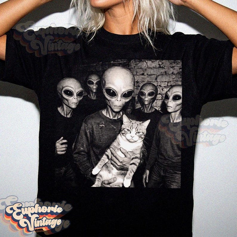 May include: Black t-shirt featuring a grayscale image of several aliens with large black eyes, one holding a cat. The image has a vintage aesthetic. The text "Euphorie Vintage" is printed on the shirt.