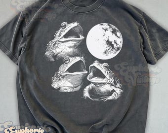 May include: A charcoal gray short-sleeve t-shirt featuring a white graphic print of three toads with their mouths open, positioned beneath a large, detailed full moon. The shirt has a crew neckline and a faded, washed-out fabric appearance.