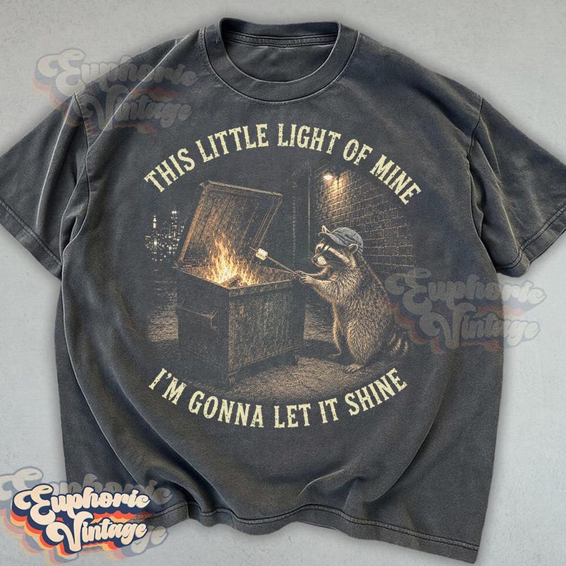 May include: A charcoal gray short-sleeve t-shirt featuring a graphic of a raccoon wearing a cap, roasting a marshmallow over a fire in an open dumpster. The text reads, "THIS LITTLE LIGHT OF MINE I'M GONNA LET IT SHINE" in a circular beige font.