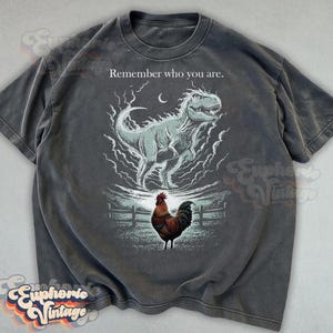 May include: Dark gray t-shirt with a graphic of a Tyrannosaurus Rex above a rooster, with the text "Remember who you are." The dinosaur is light blue and surrounded by white clouds. The rooster is brown and red.