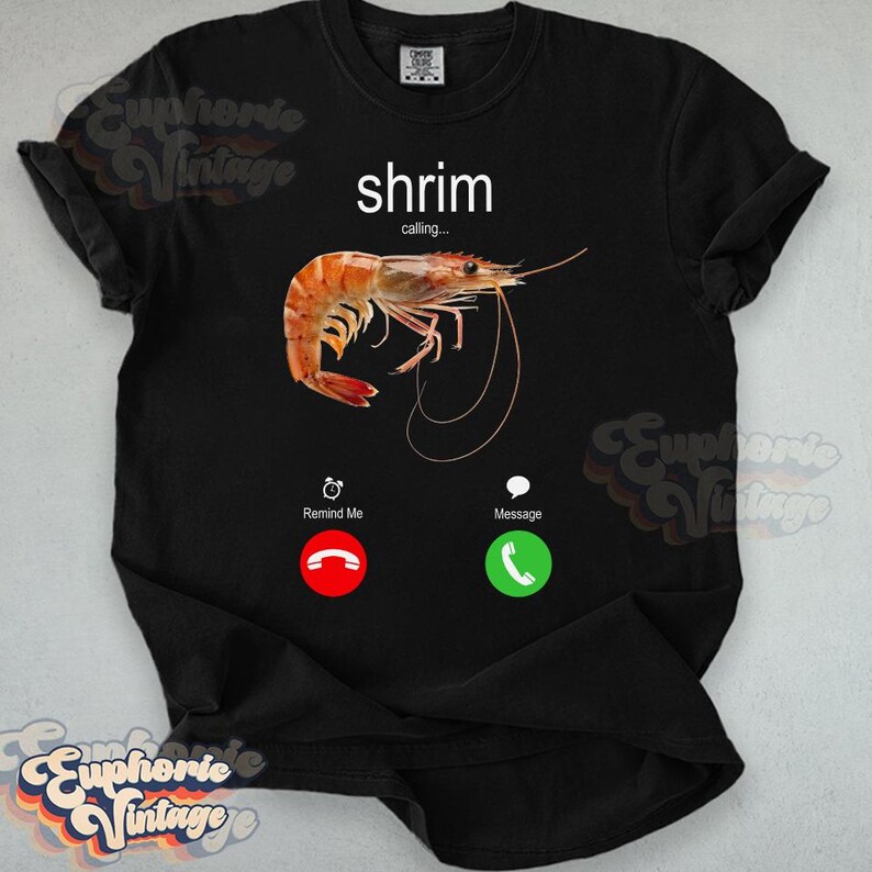 Op de afbeelding: Zwart t-shirt met een afbeelding van een garnaal en het woord "shrimp" erboven, met "calling..." eronder. Het ontwerp bevat een rode "Remind Me"-knop en een groene "Message"-knop.