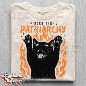 May include: Off-white crew neck t-shirt with a black cat illustration and orange flames. The text "BURN THE PATRIARCHY" is printed above the cat. The shirt has a vintage aesthetic.