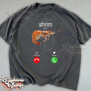 Op de afbeelding: Donkergrijs t-shirt met een garnalenafbeelding en de tekst "shrim calling...". Het ontwerp bevat een rode weigerknop en een groene accepteerknop. Het t-shirt heeft een vintage uitstraling.