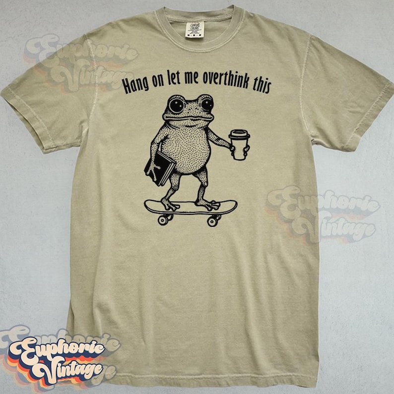Puede incluir: Una camiseta beige de manga corta con un gr&aacute;fico negro de una rana en un monopat&iacute;n sosteniendo un libro y una taza de caf&eacute;. Sobre la rana, el texto dice, Hang on let me overthink this. La camiseta tiene un cuello redondo est&aacute;ndar.