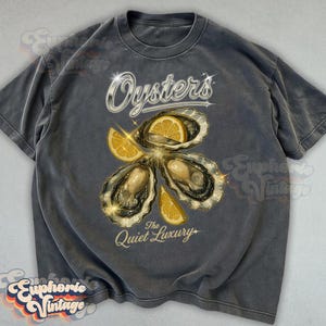 May include: Gray t-shirt with an oyster graphic. The design features three oysters with lemon slices, the word "Oysters" in a silver font, and the phrase "The Quiet Luxury". The shirt has a vintage aesthetic.