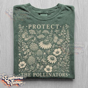 May include: A forest green crew neck t-shirt featuring a cream-colored graphic print of various wildflowers, butterflies, and bees. The text PROTECT THE POLLINATORS is printed in a serif font above and below the detailed botanical illustration.