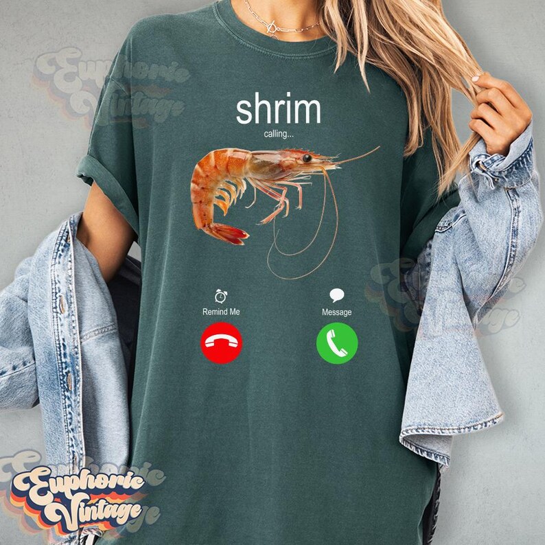 Op de afbeelding: Donkergroen t-shirt met een garnalenafbeelding en het woord "shrimp" erboven. De afbeelding lijkt op een telefoonscherm met een garnalenafbeelding, een rode "Remind Me"-knop en een groene "Message"-knop.