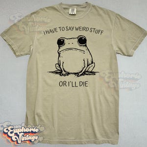 May include: A tan t-shirt with a black illustration of a frog and the text "I HAVE TO SAY WEIRD STUFF OR I'LL DIE". The frog has large eyes and a grumpy expression. The shirt has short sleeves and a crew neck.