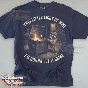 May include: A navy blue t-shirt featuring a graphic of a raccoon wearing a cap, standing by a dumpster fire and roasting a marshmallow on a stick. The text reads, "THIS LITTLE LIGHT OF MINE" above and "I'M GONNA LET IT SHINE" below the graphic.