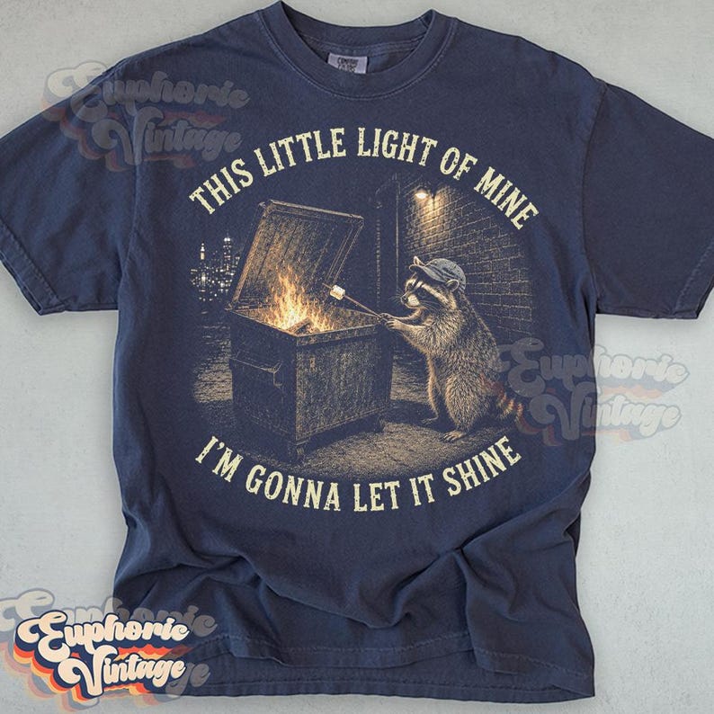 May include: A navy blue t-shirt featuring a graphic of a raccoon wearing a cap, standing by a dumpster fire and roasting a marshmallow on a stick. The text reads, "THIS LITTLE LIGHT OF MINE" above and "I'M GONNA LET IT SHINE" below the graphic.
