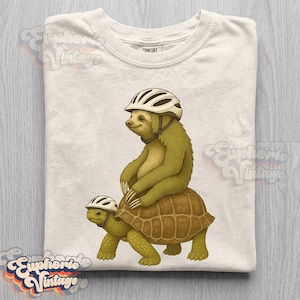 May include: A cream-colored t-shirt featuring a graphic illustration of a sloth wearing a white bicycle helmet riding on the back of a tortoise that is also wearing a white bicycle helmet. The shirt has a crew neckline and short sleeves.