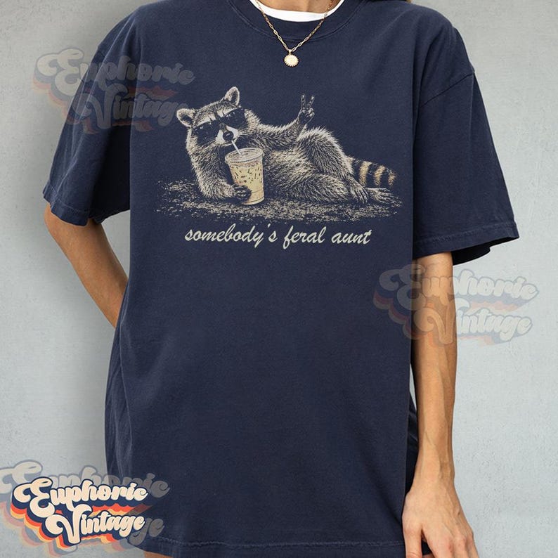 May include: A navy blue short-sleeve t-shirt featuring a graphic of a raccoon wearing sunglasses, holding a drink, and making a peace sign. Below the raccoon, the text reads, "somebody's feral aunt" in a white, lowercase, cursive-style font.