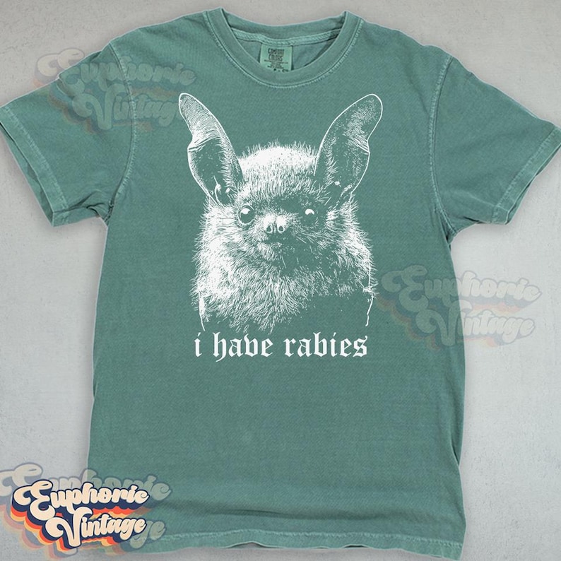 May include: A teal-colored t-shirt featuring a white graphic of a bat's face with large ears. Below the bat, the text reads "i have rabies" in a gothic font. The shirt has a crew neck and short sleeves.