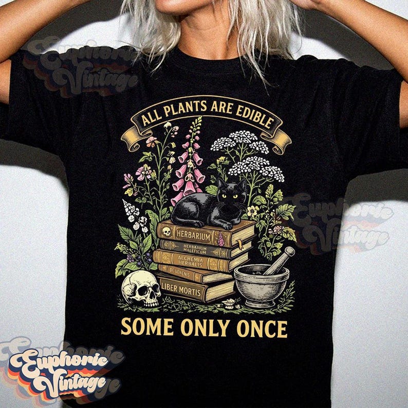 May include: A black t-shirt featuring a graphic print of a black cat resting on a stack of books, surrounded by botanical illustrations, a human skull, and a mortar and pestle. The text reads ALL PLANTS ARE EDIBLE at the top and SOME ONLY ONCE below.