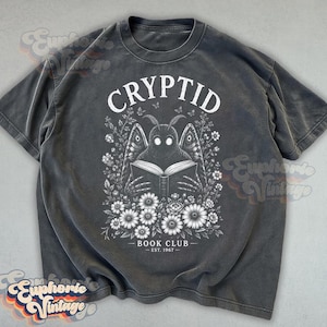 May include: A charcoal gray short-sleeve t-shirt featuring a white graphic print of a moth-like creature reading an open book. The design includes floral elements, small butterflies, and the text "CRYPTID BOOK CLUB EST. 1967" in a white font.
