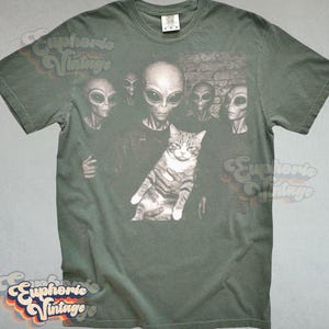 May include: Green t-shirt featuring a graphic of several aliens surrounding a cat. The aliens have large black eyes and gray skin. The cat is gray and white. The shirt has a vintage look.