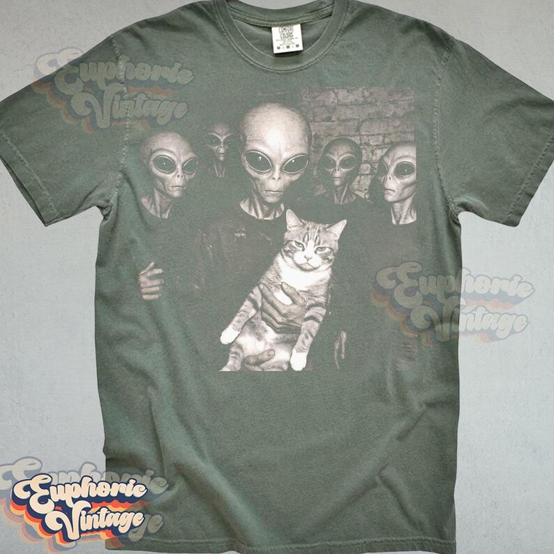 May include: Green t-shirt featuring a graphic of several aliens surrounding a cat. The aliens have large black eyes and gray skin. The cat is gray and white. The shirt has a vintage look.