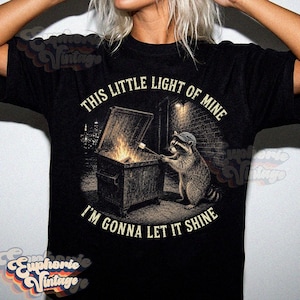 May include: A black t-shirt featuring a graphic of a raccoon wearing a cap, standing by a dumpster fire and roasting a marshmallow on a stick. The text "THIS LITTLE LIGHT OF MINE" and "I'M GONNA LET IT SHINE" is printed in a circular arc around the image.