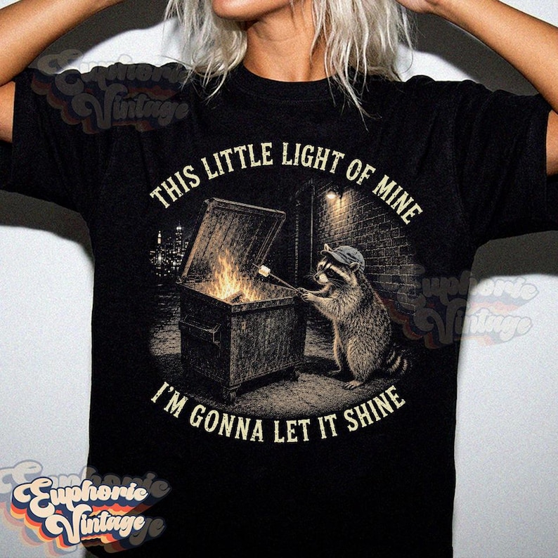 May include: A black t-shirt featuring a graphic of a raccoon wearing a cap, standing by a dumpster fire and roasting a marshmallow on a stick. The text "THIS LITTLE LIGHT OF MINE" and "I'M GONNA LET IT SHINE" is printed in a circular arc around the image.