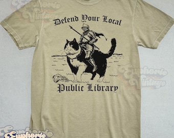 May include: Tan t-shirt with a graphic of a knight riding a large black and white cat, holding a spear. The text "Defend Your Local Public Library" is printed above and below the image in a medieval font.
