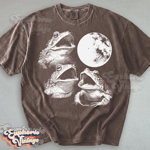 May include: Brown t-shirt featuring a graphic of three frogs with open mouths and a full moon. The design is in white, creating a contrast against the shirt's color. The shirt has a vintage aesthetic.