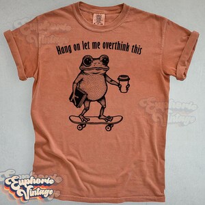 Puede incluir: Una camiseta de manga corta color terracota con un gr&aacute;fico negro de una rana en un monopat&iacute;n sosteniendo un libro y una taza de caf&eacute;. El texto sobre la rana dice, Hang on let me overthink this. La camiseta tiene un cuello redondo cl&aacute;sico.